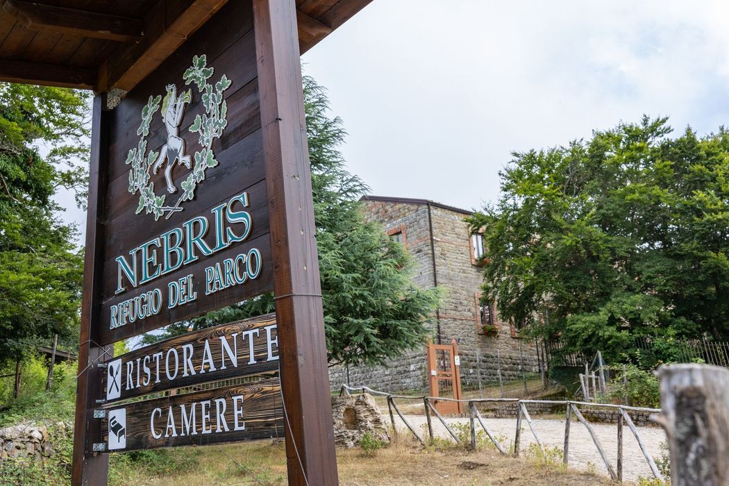 Insegna in legno con la scritta “Neris Restaurant Camere” accanto a un edificio in pietra e agli alberi, all’aperto