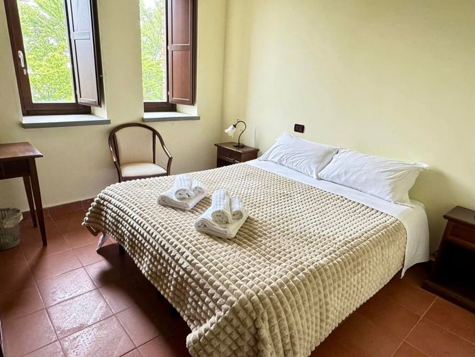 Accogliente camera da letto con un letto matrimoniale, cuscini bianchi, coperta di maglia beige e due finestre da cui entra la luce del giorno.