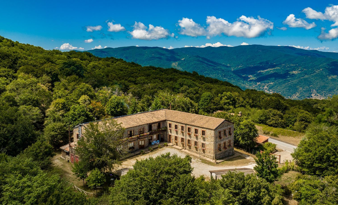 Edificio in pietra sulla cima di una collina, circondato da una rigogliosa foresta e affacciato su montagne blu in lontananza, sotto un cielo luminoso.