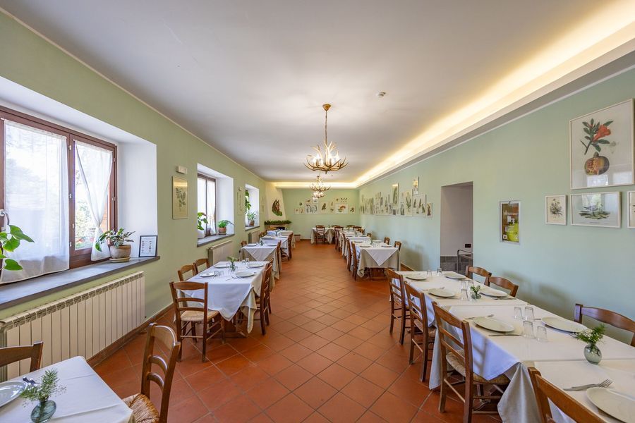 Luminosa sala da pranzo con lunghi tavoli coperti da tovaglie bianche, pavimento in terracotta e finestre lungo la parete sinistra.