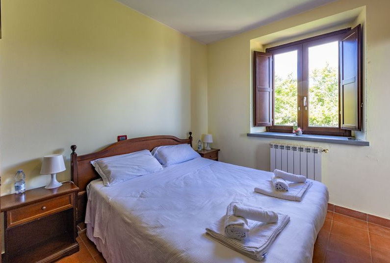 Camera da letto piccola con un letto matrimoniale, un comodino e una finestra aperta da cui entra la luce del giorno.
