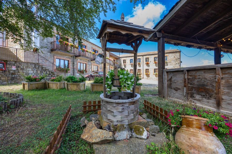Pozzo in pietra in un cortile rustico con fiori, erba e antichi edifici in pietra sotto un cielo azzurro