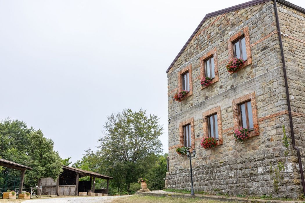 Casale in pietra con persiane rosse in un cortile di campagna, alberi e ripari rustici nelle vicinanze.