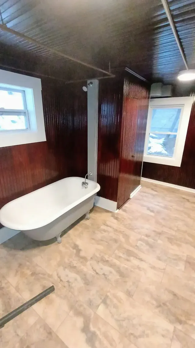 Small bathroom with a white bathtub, dark wood-paneled walls, wood floor, and a window letting in daylight.