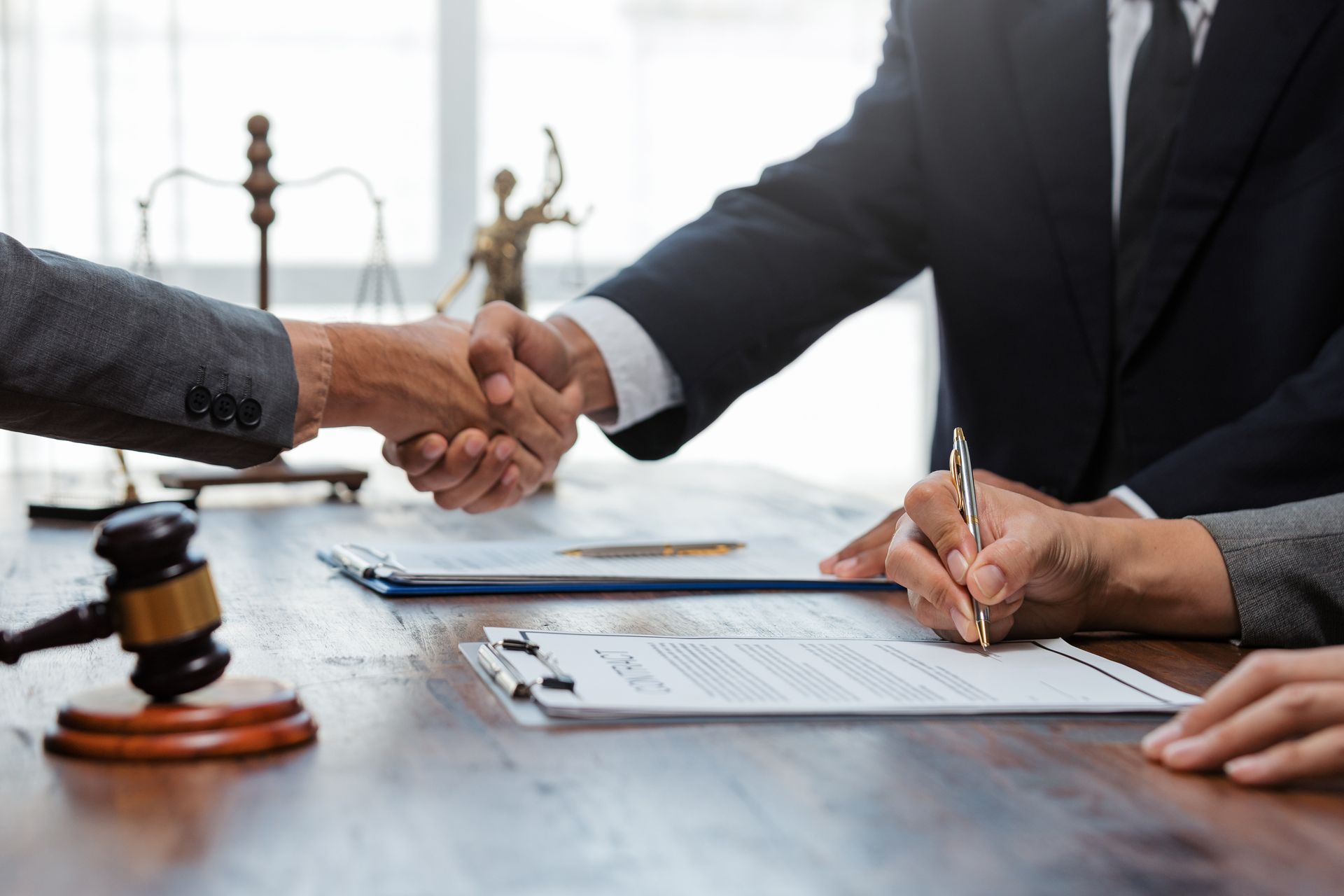 A lawyer and a client shaking hands across a desk, with another lawyer signing a document.