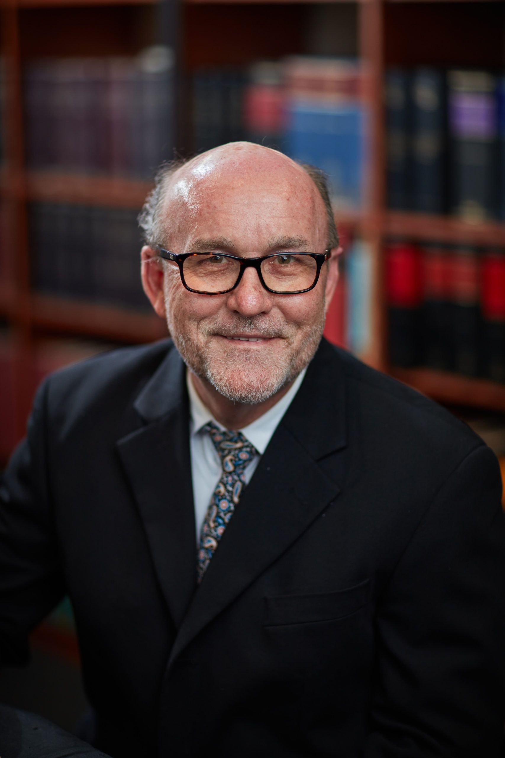 A man in a suit and tie is sitting in front of a bookshelf.