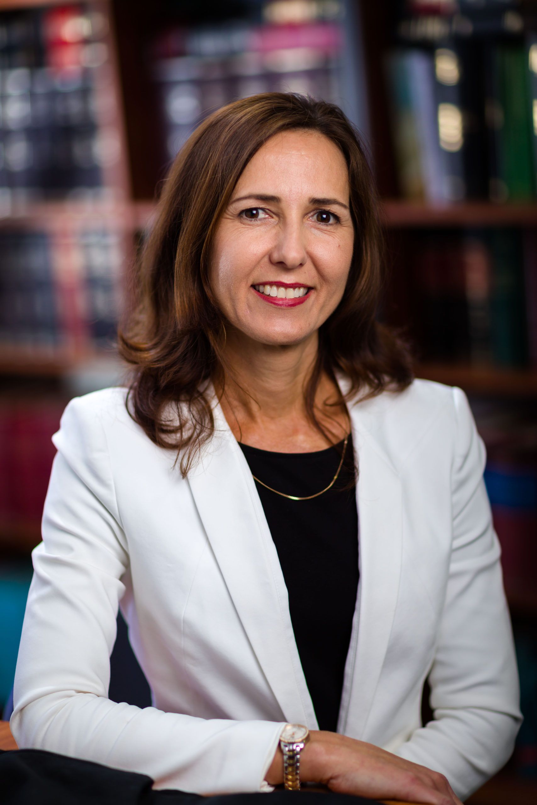 A woman in a white jacket is sitting at a table in front of a bookshelf.