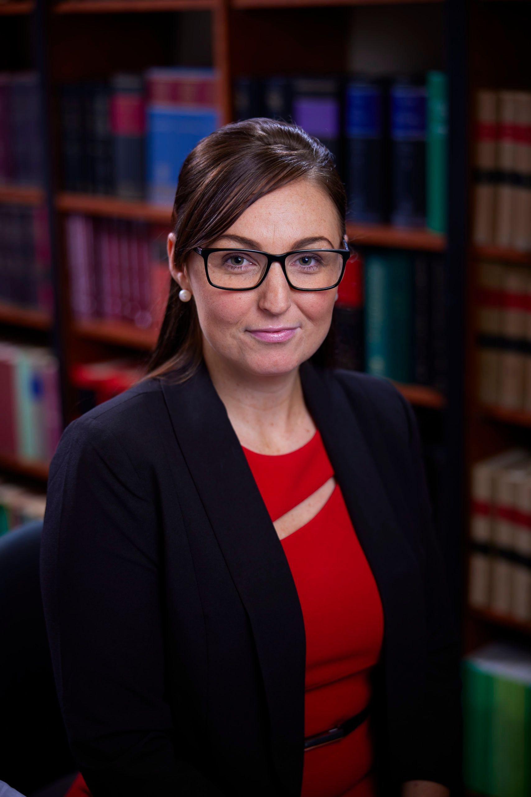A woman wearing glasses and a red dress is sitting in front of a bookshelf.
