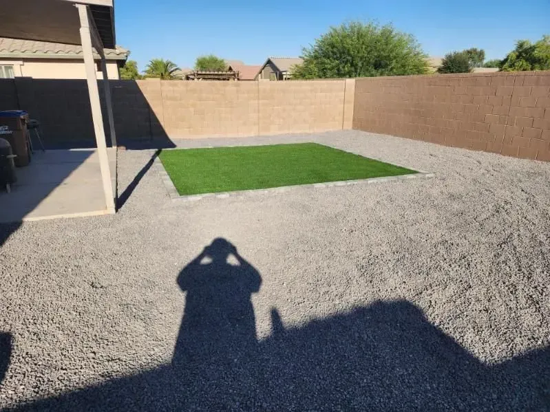 Gravel backyard with a small green lawn patch, beige walls, and a covered patio on the left
