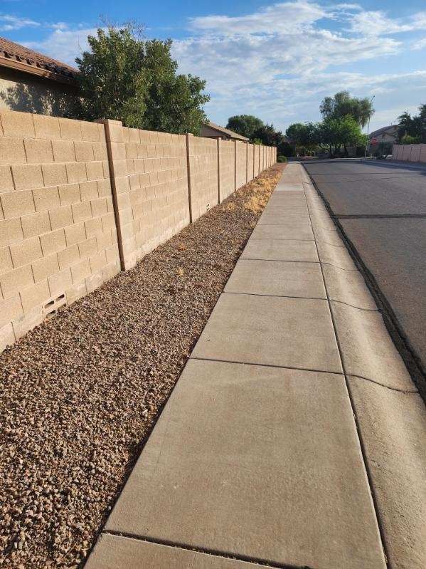 Residential sidewalk beside tan block wall and gravel strip along a quiet street under a blue sky