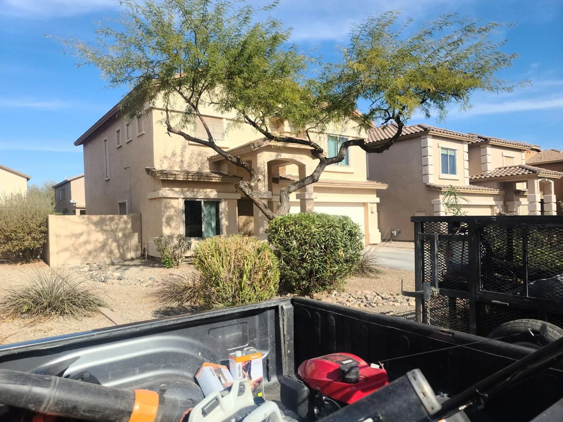 Desert-style house with trees and shrubs, viewed over a pickup truck bed loaded with tools and gear.
