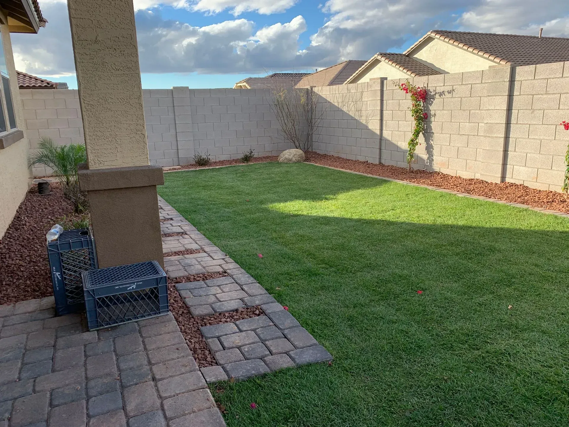 Backyard patio with pavers, green lawn, stone walls, and a blue chair under a covered post.