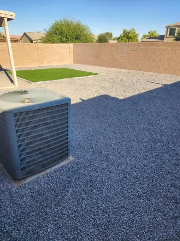Backyard with gravel ground, tan fence, patch of grass, and an outdoor air conditioner unit.
