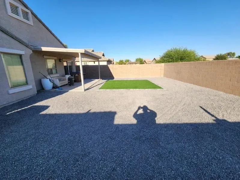 Backyard with gravel ground, covered patio, tan privacy wall, and small green lawn patch under a clear blue sky