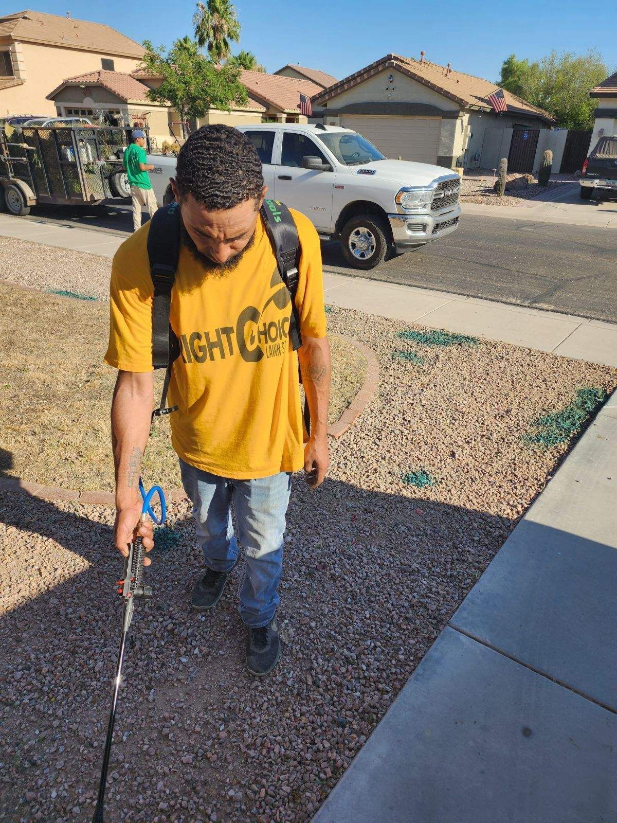 Person walking on a gravel driveway with a cane, wearing a yellow shirt and jeans in a suburban neighborhood.