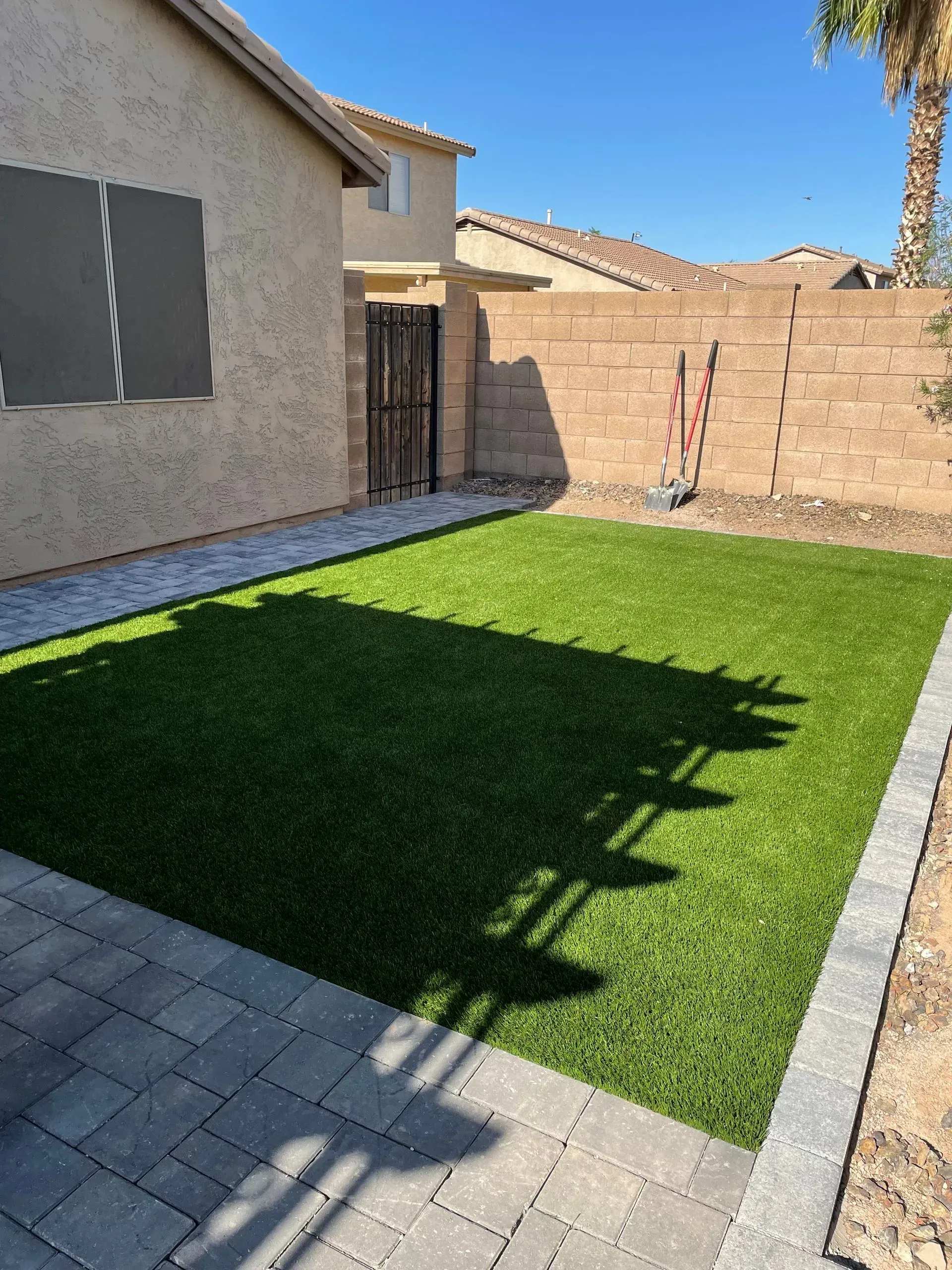 Small backyard patio with artificial grass, paved border, stucco house wall, and brick fence in sunlight