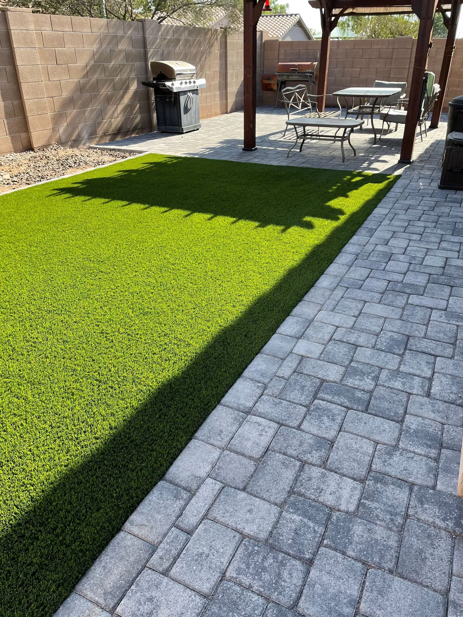 Backyard patio with green lawn, stone pavers, grill, and outdoor dining set in sunlight