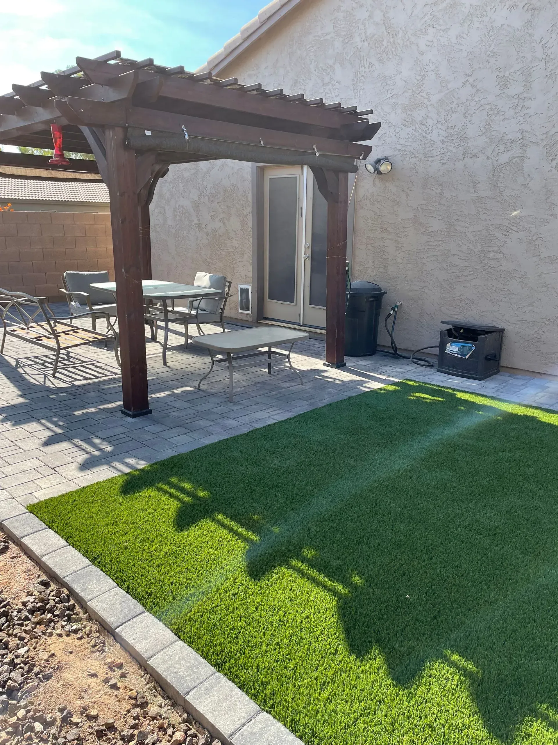 Backyard patio with a wooden pergola, table and chairs, and green artificial turf beside a stucco wall