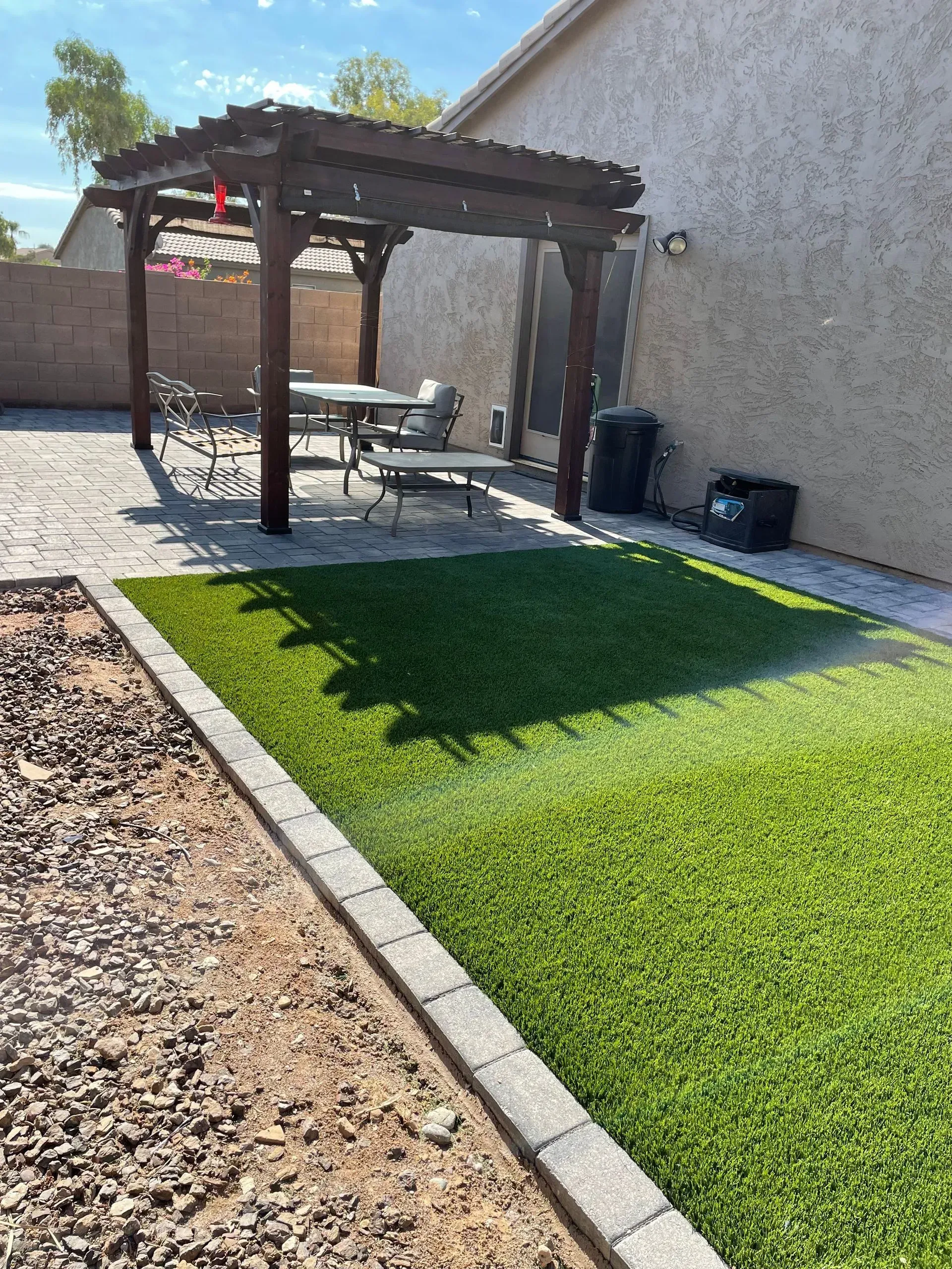 Backyard patio with a wooden pergola, picnic table, and green artificial turf beside a gravel path.