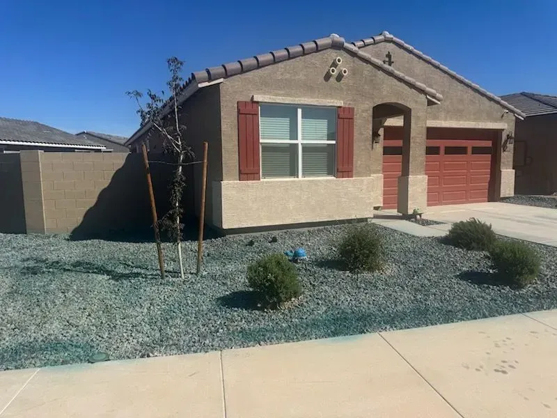 Single-story beige house with red shutters and garage, gravel front yard, and young trees under a blue sky