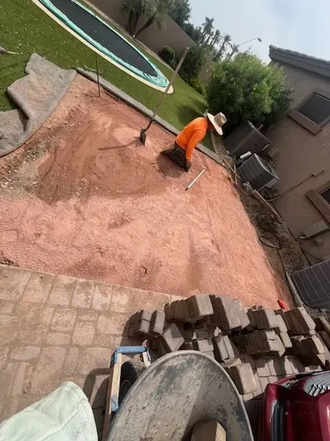 Worker in orange shirt leveling red dirt in a backyard construction area.