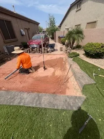 Person in orange shirt working in a red dirt excavation in a residential backyard with a parked car nearby