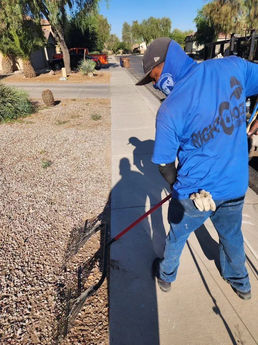 Worker in a blue hoodie using a trimmer along a sidewalk beside gravel and trees.