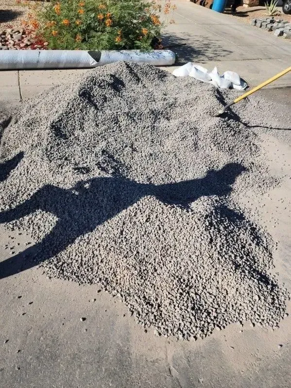 Pile of gray gravel on a driveway beside a yellow hose and garden bed, with a long shadow across it