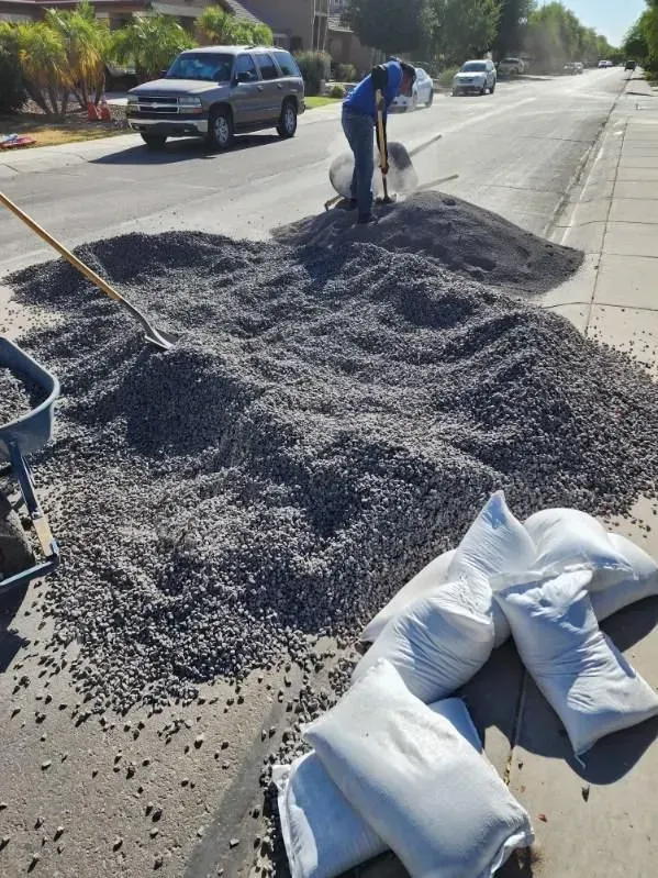 Worker shoveling gravel from a roadside pile beside white sandbags on a sunny street