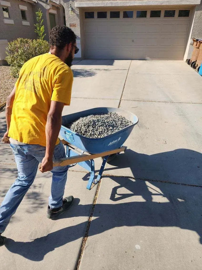 Person pushing a wheelbarrow of rocks up a driveway toward a closed garage door