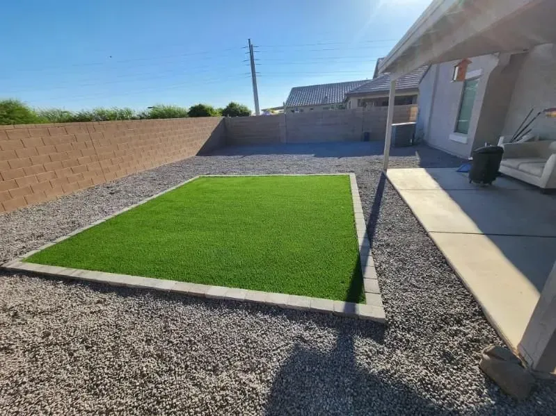 Small landscaped backyard with green artificial turf, gravel border, and a covered patio beside a tan wall.