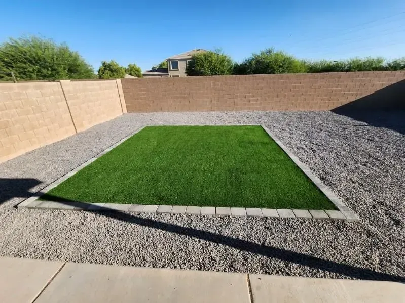 Rectangular green lawn in a gravel courtyard, bordered by concrete and tan walls under a clear blue sky