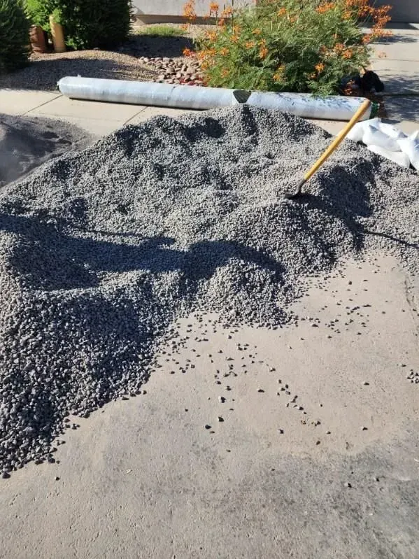 Pile of black gravel spilled on a driveway with a broom nearby and a rolled white barrier in the background