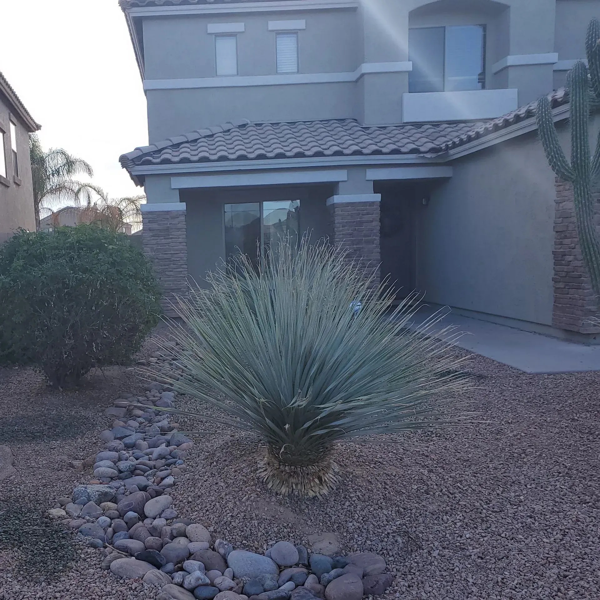 Modern two-story stucco house with desert landscaping, gravel yard, river rocks, and a spiky agave plant in front.