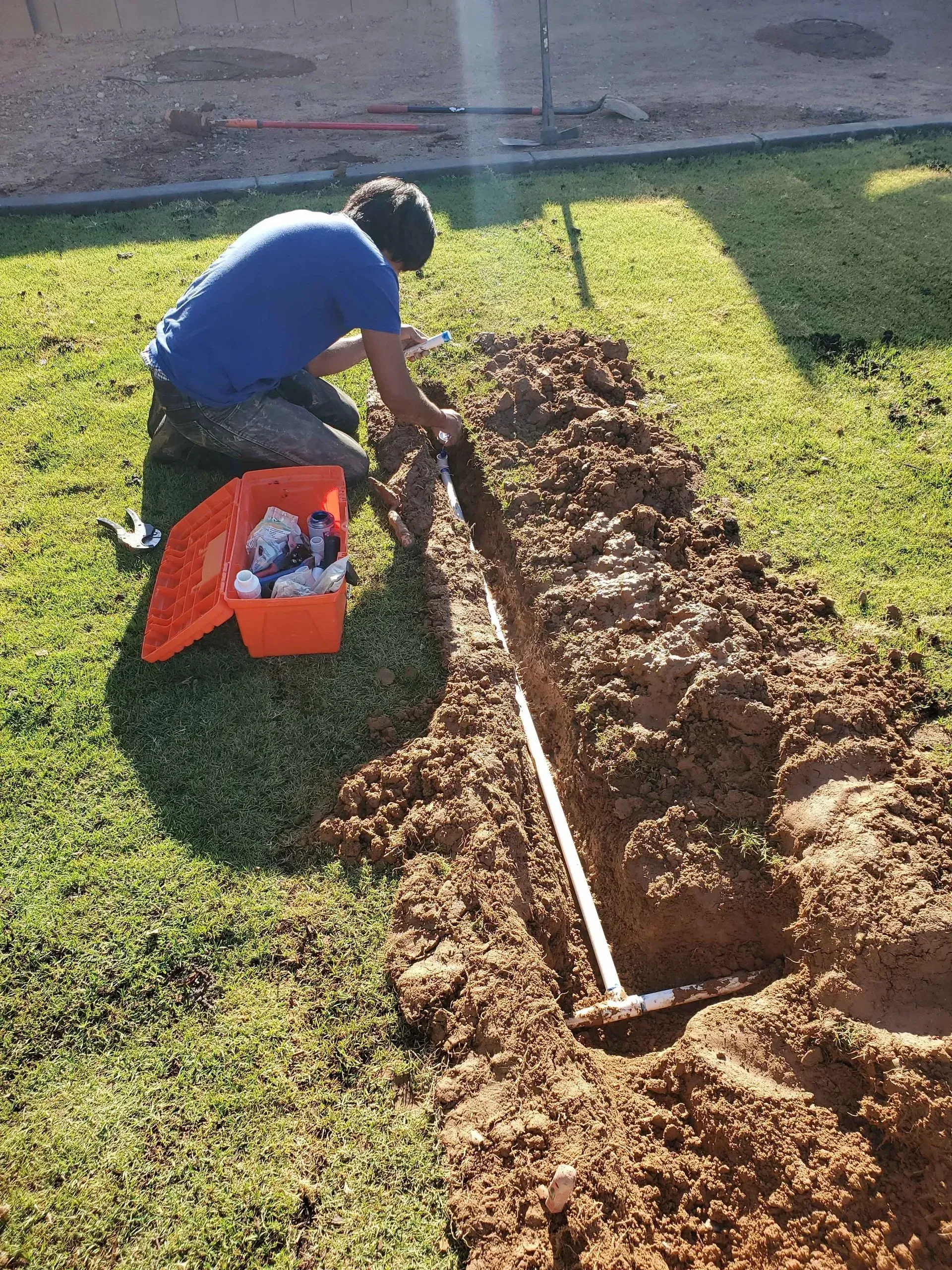 Person digging a garden bed with a rake beside an orange crate on a sunny lawn
