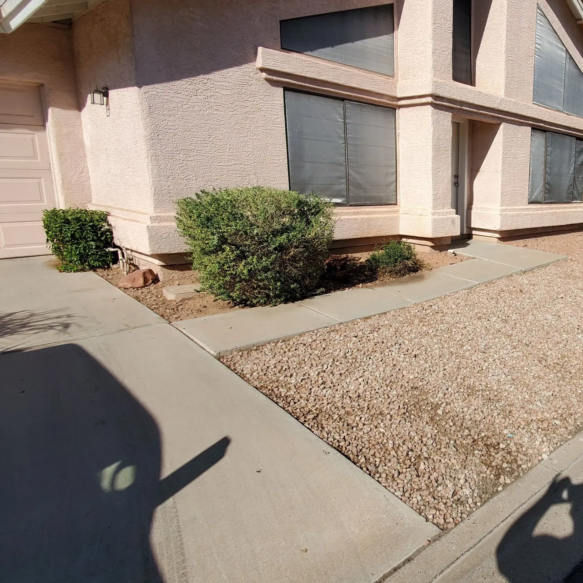 Front of a beige stucco house with windows, shrubs, a sidewalk, and gravel landscaping in bright sunlight
