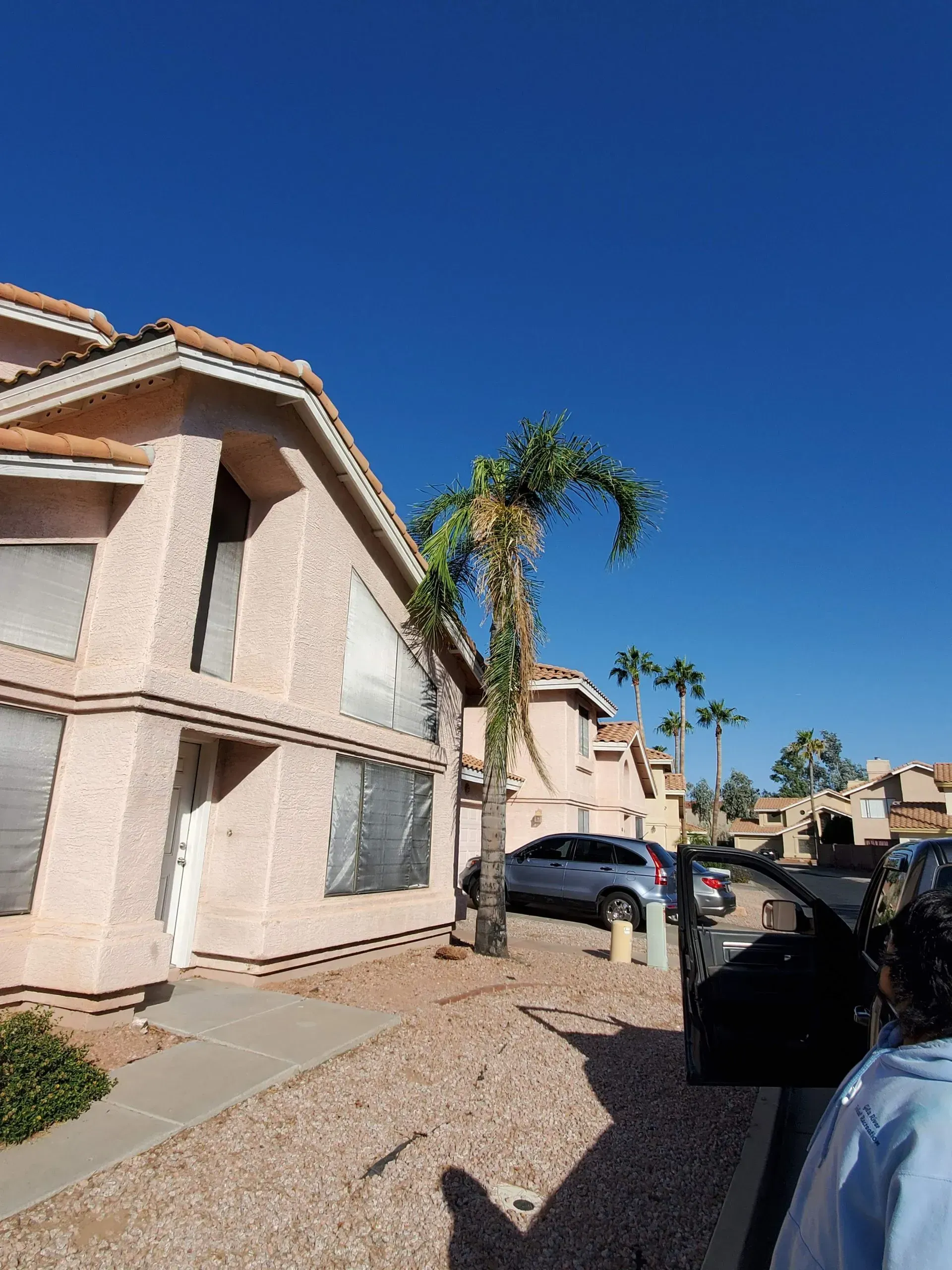Suburban townhouse row with parked cars, gravel driveway, and palm trees under a clear blue sky