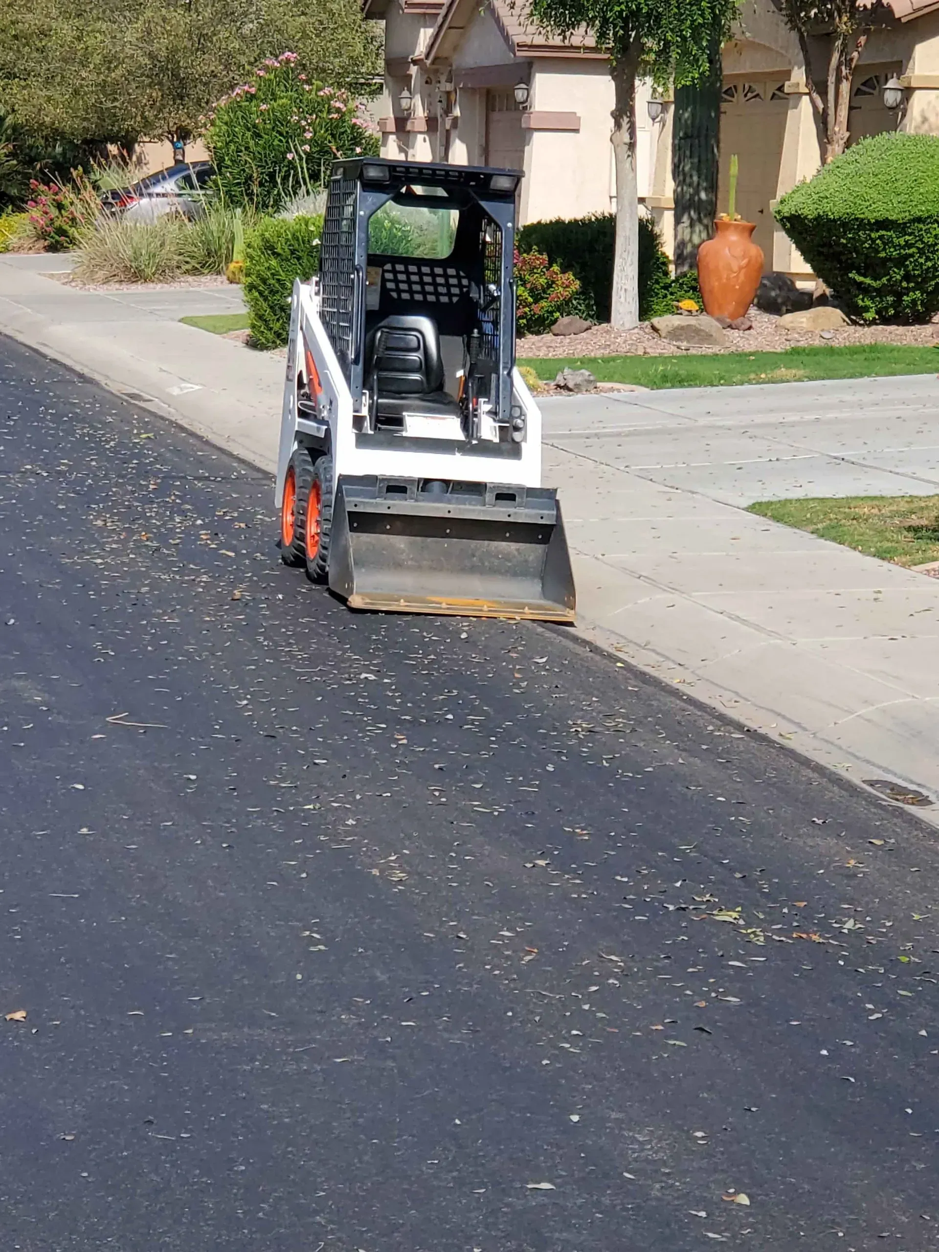 Compact skid-steer with broom attachment cleaning debris along a residential curbside street