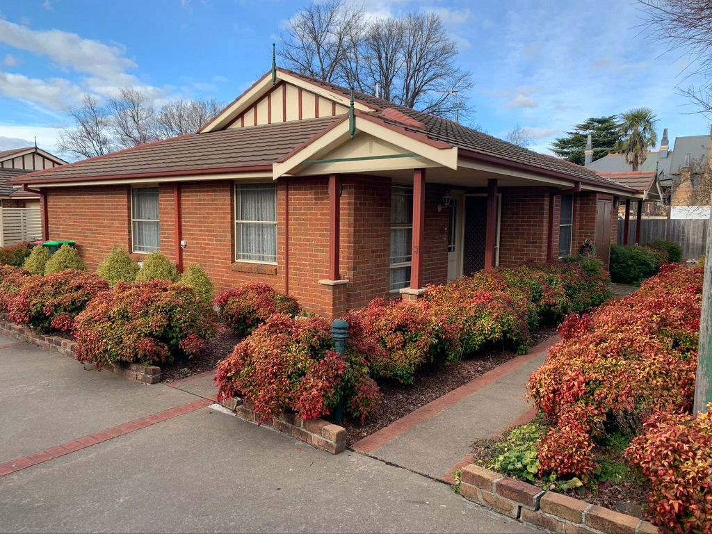 Red brick single-story house with red and green bushes in front, path, and a blue sky. — Tamworth Strata Management Services in Armidale, NSW