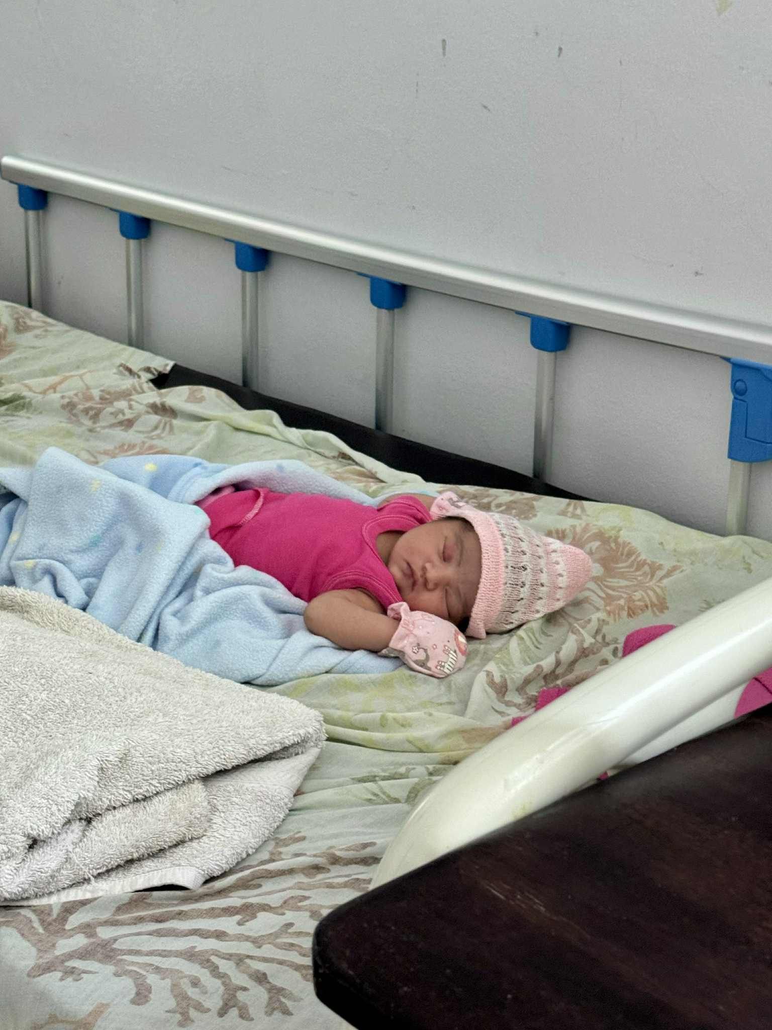 Newborn sleeping on a hospital bed, wearing a pink shirt and hat, covered with a blue blanket.
