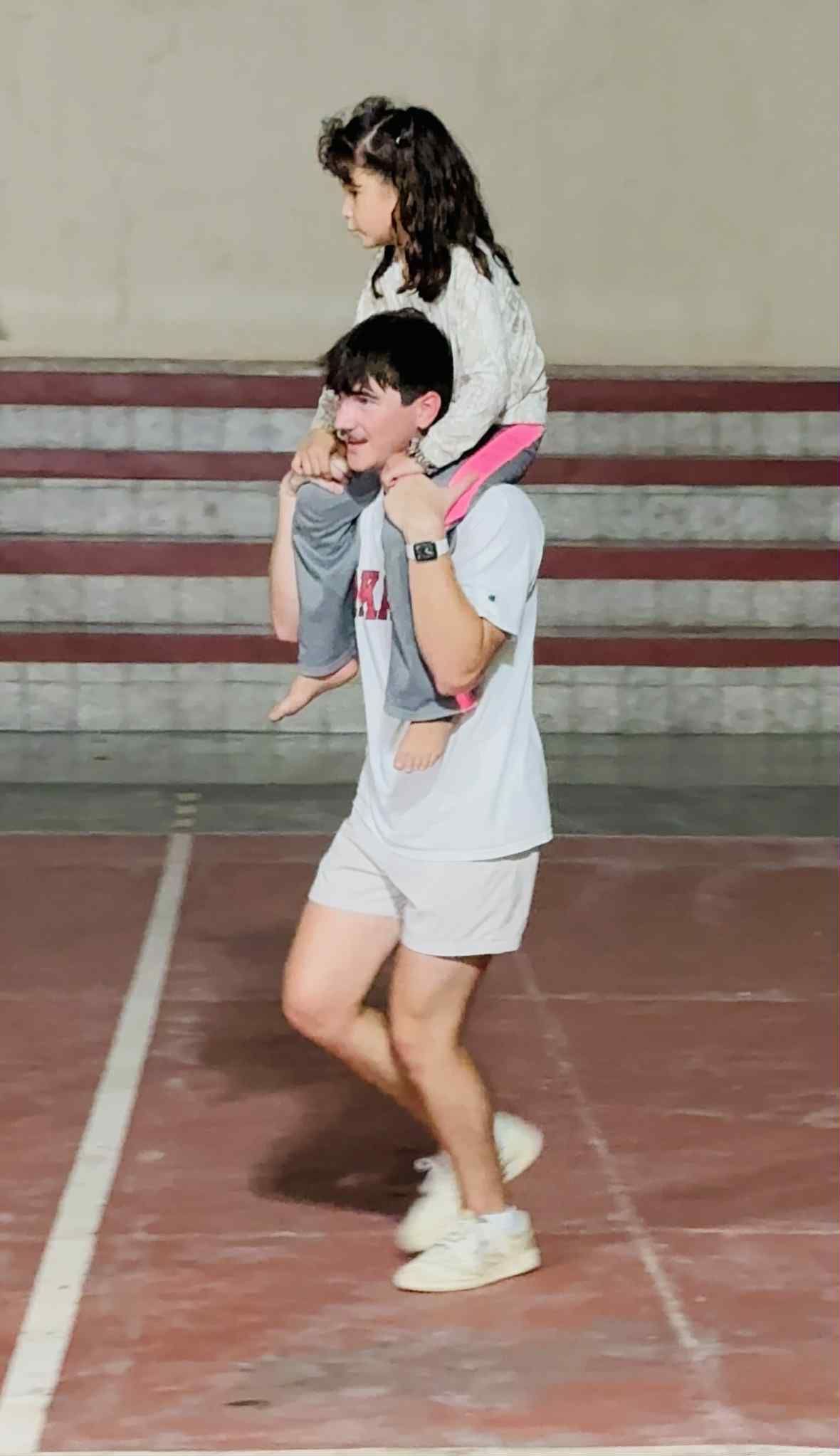 Young person carrying a child on their shoulders on a red floor, possibly an indoor court.