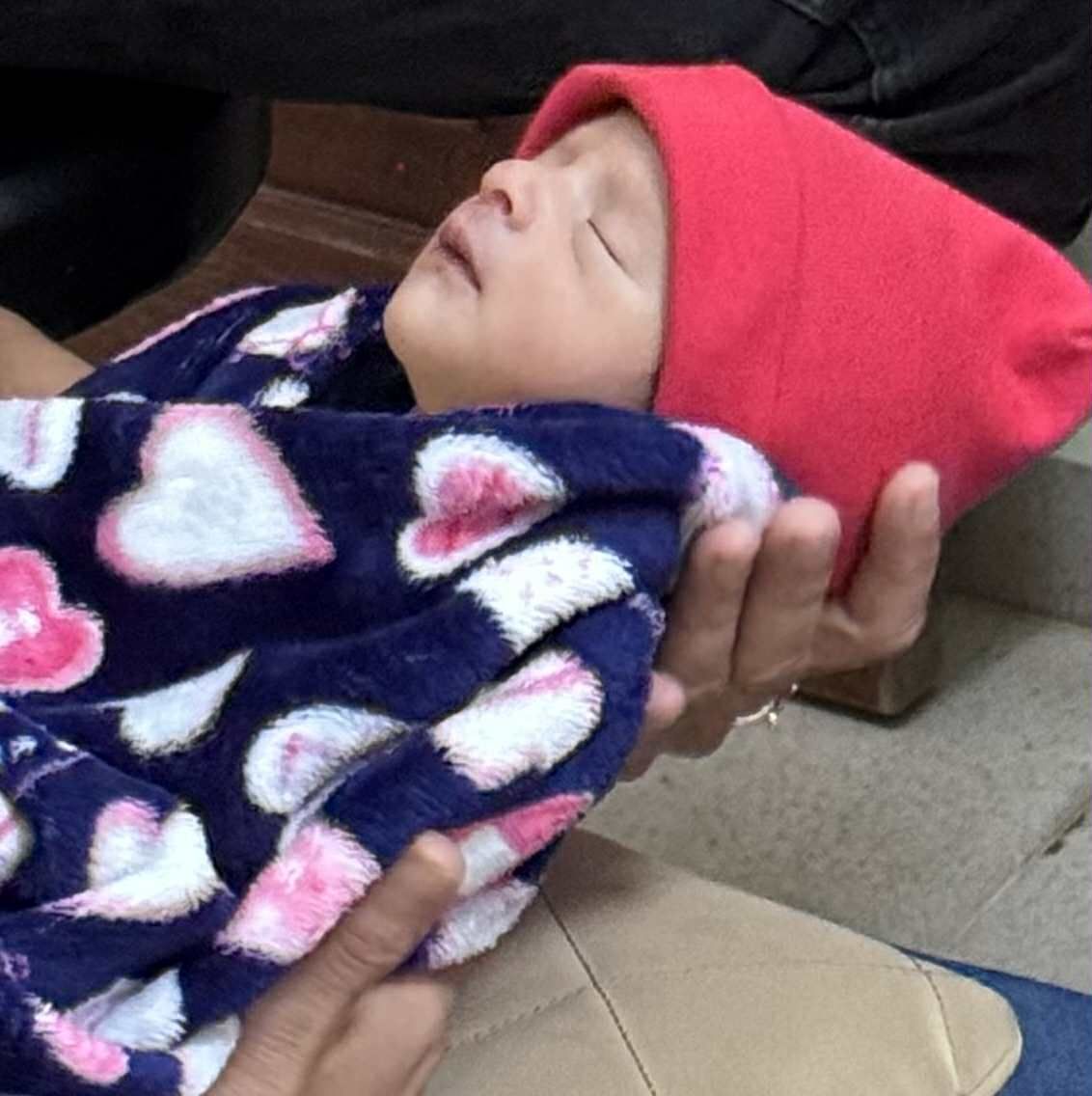 Sleeping baby in a red hat, wrapped in a blue blanket with pink hearts, held by someone.