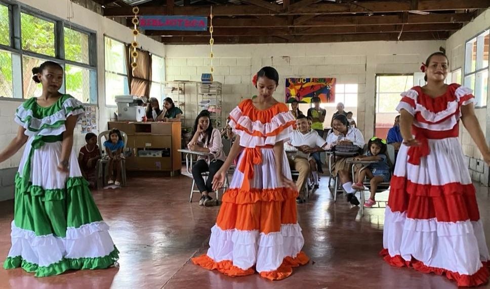 Three dancers in colorful ruffled dresses perform in a room, with an audience watching.