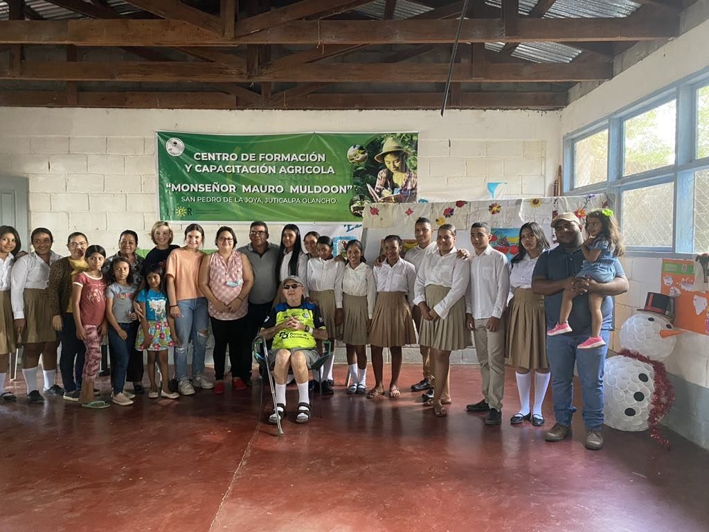 Group of people posing together inside a room. Banner in the background. Several children wearing school uniforms.