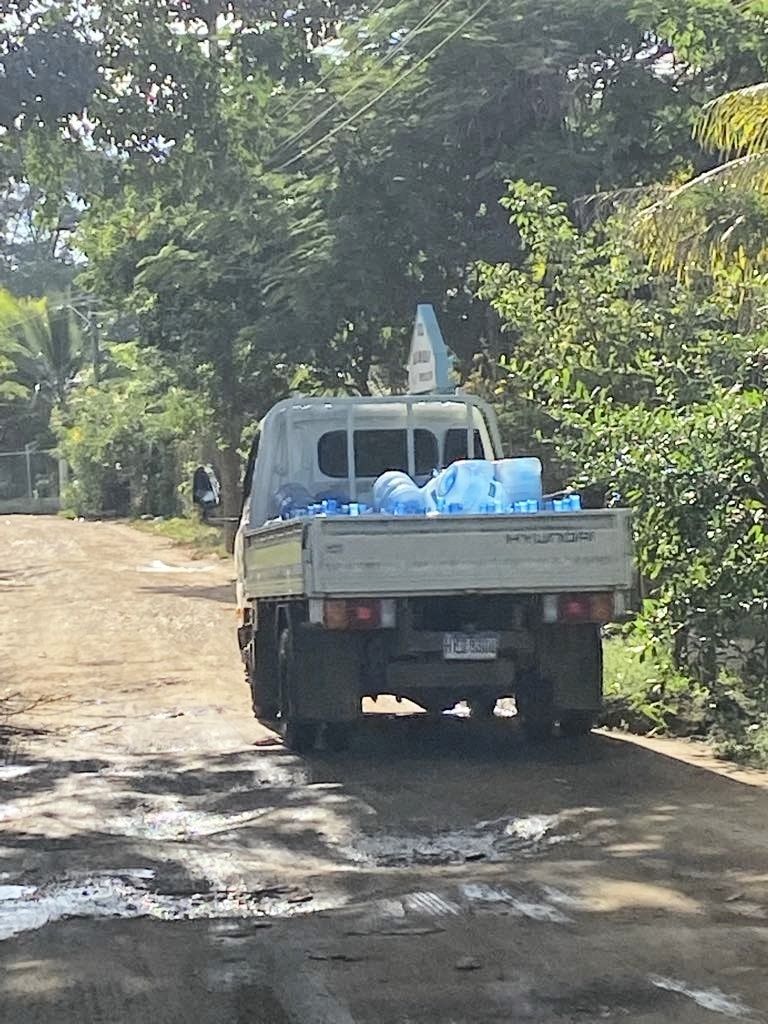 White Hyundai truck carrying blue water jugs on a dirt road. Trees line the sides.