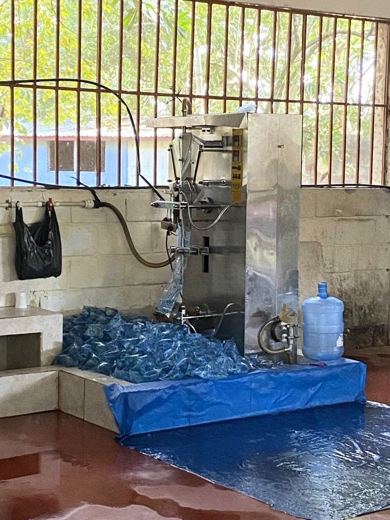 A water sachet packaging machine with filled sachets on a blue tarp floor, located near a window.