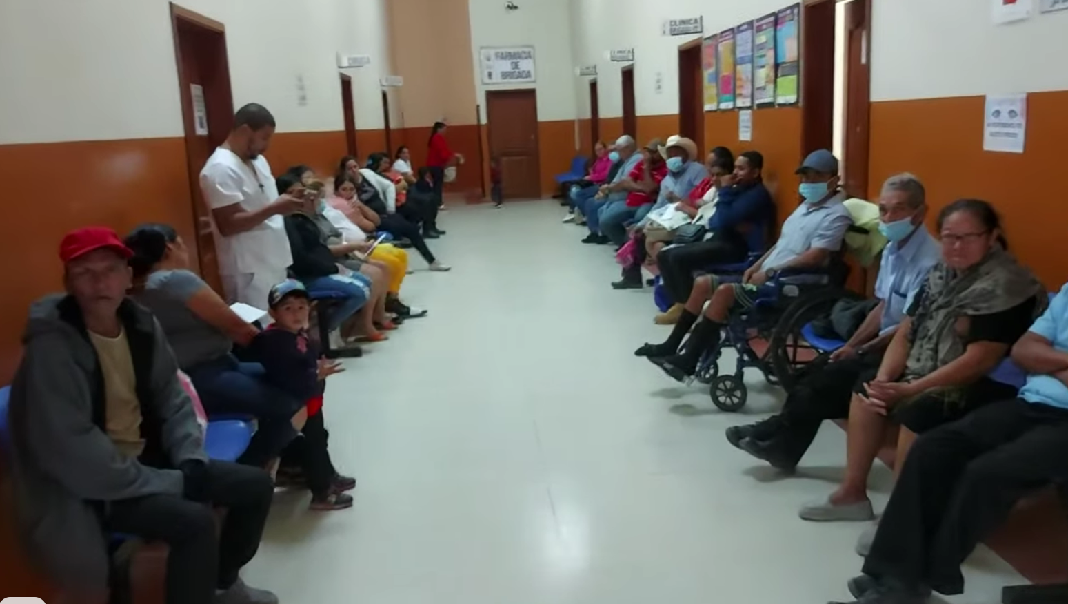 People waiting in a hospital waiting room, some wearing masks. Several are seated on benches.