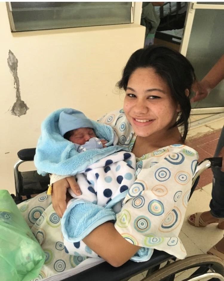 Woman in wheelchair holds newborn, wrapped in blue blanket; hospital setting.