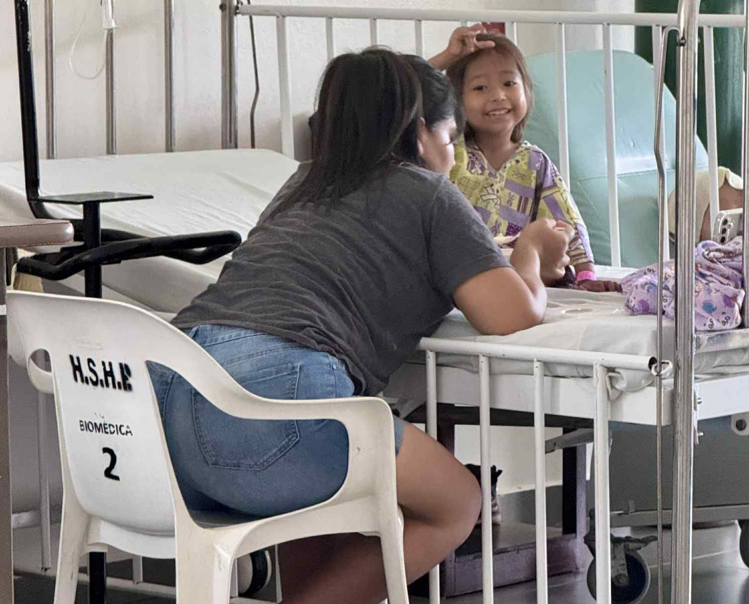Woman sits with a child in hospital. The child smiles while sitting on a bed.