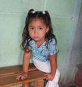 Girl leaning on a wooden bench, wearing a blue shirt and white pants, with pigtails. Light green background.
